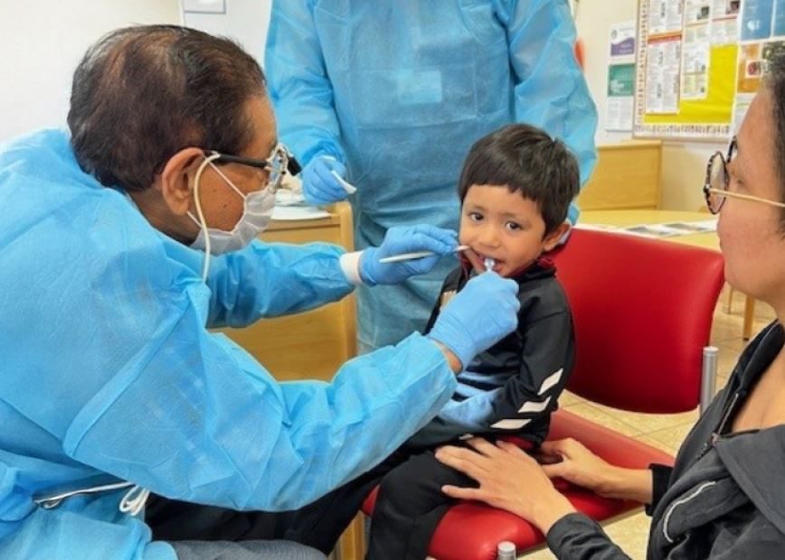 Un dentista con bata y mascarilla azules examina los dientes de un niño sentado en una silla roja. Una mujer, posiblemente su tutora, le sujeta las manos, y otra persona con equipo de protección permanece cerca.