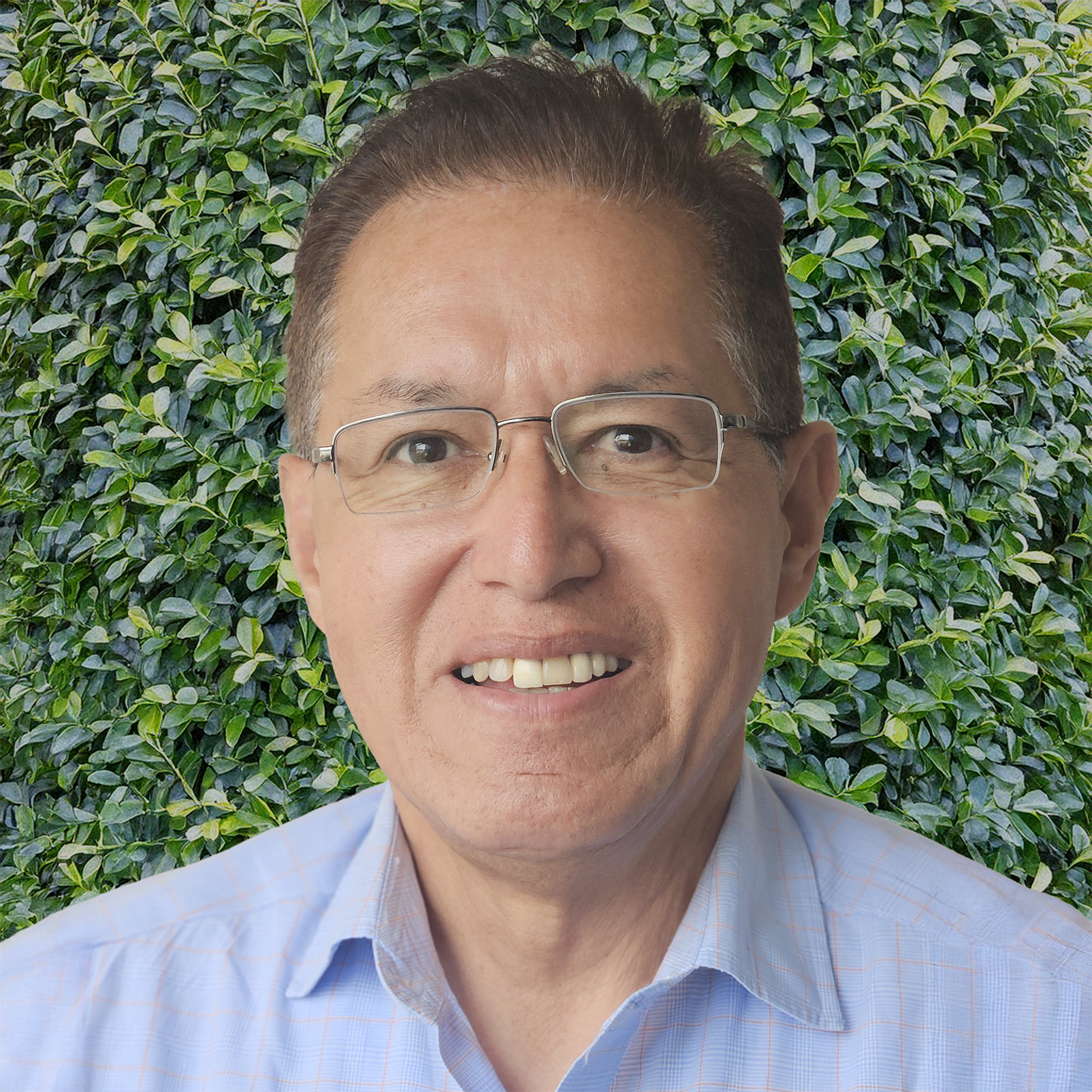 Un hombre con gafas y camisa de cuadros azul claro sonríe delante de un exuberante fondo de hojas verdes.