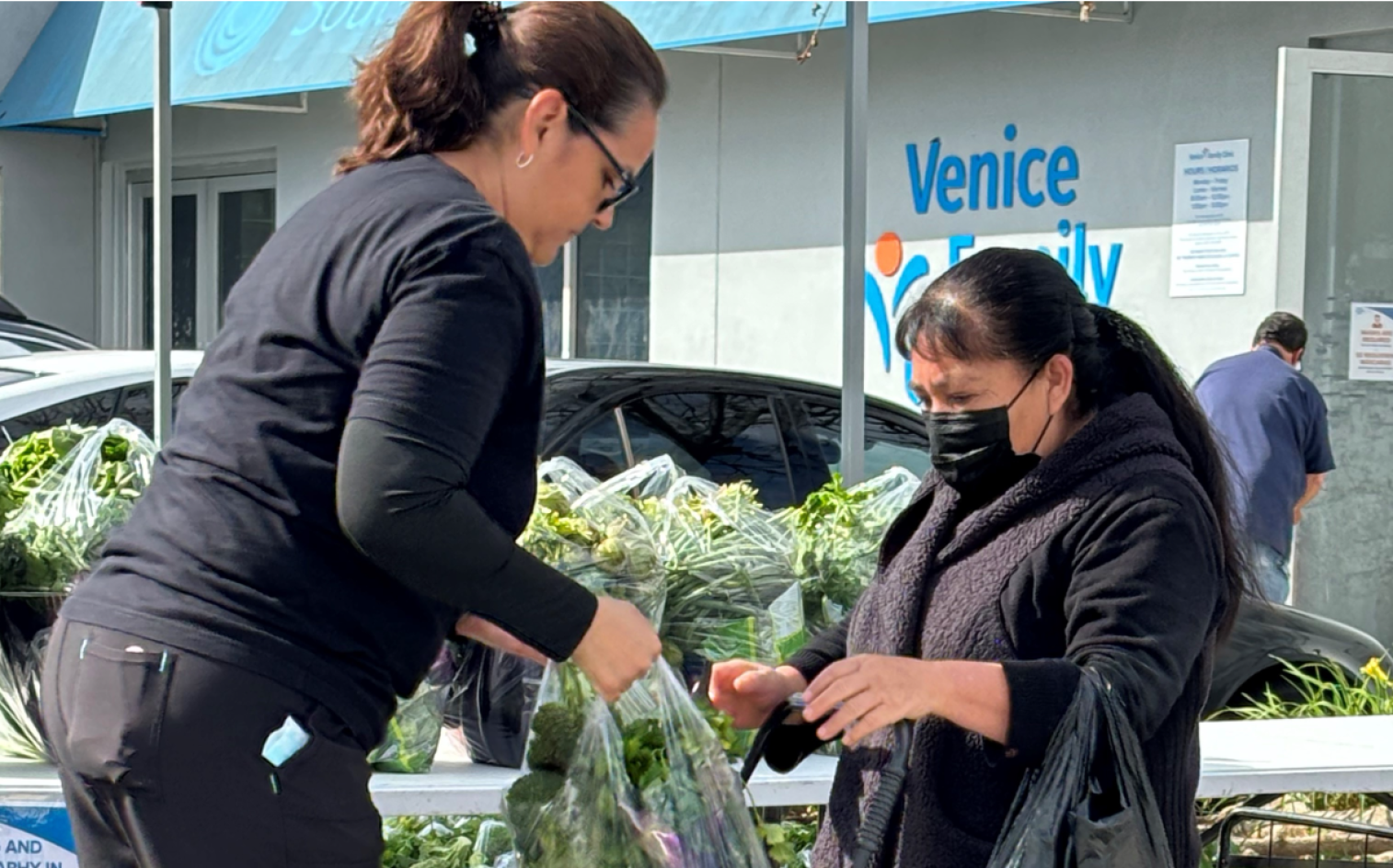 A Woman Hands A Bag Of Leafy Greens To Another Woman Wearing A Mask And Holding A Reusable Shopping Bag At An Outdoor Market Stand. A Building With A “Venice Family” Sign Is Visible In The Background. A Woman Hands A Bag Of Leafy Greens To Another Woman Wearing A Mask And Holding A Reusable Shopping Bag At An Outdoor Market Stand. A Building With A “Venice Family” Sign Is Visible In The Background.