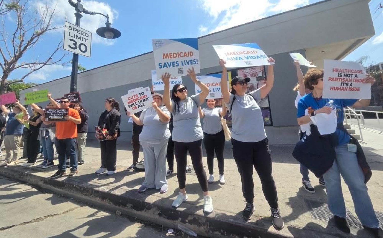A Group Of People Stand On A Sidewalk Holding Signs In Support Of Medicaid And Healthcare Access. The Signs Read Messages Such As MEDICAID SAVES LIVES! And HEALTHCARE IS NOT A PARTISAN ISSUE.