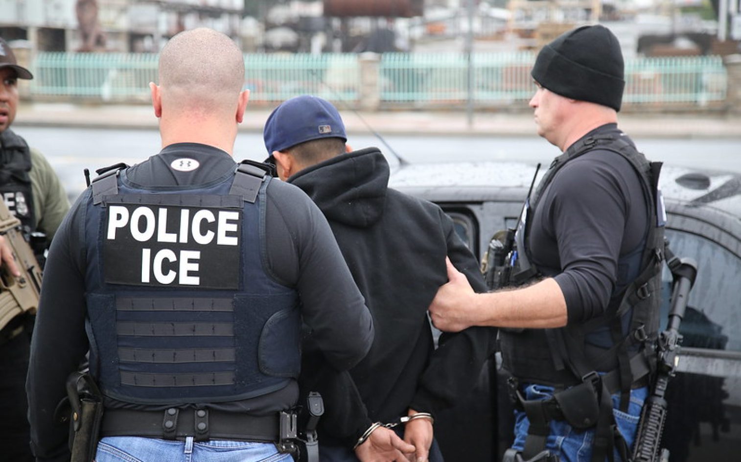 Several Law Enforcement Officers Wearing “POLICE ICE” Vests Detain A Handcuffed Person In A Black Hoodie Next To A Black Vehicle On A City Street. Several Law Enforcement Officers Wearing “POLICE ICE” Vests Detain A Handcuffed Person In A Black Hoodie Next To A Black Vehicle On A City Street.