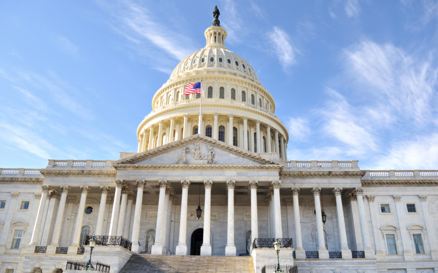 The United States Capitol Building, Featuring A Large Dome, Columns, And An American Flag Flying In Front, Set Against A Blue Sky With Wispy Clouds. The United States Capitol Building, Featuring A Large Dome, Columns, And An American Flag Flying In Front, Set Against A Blue Sky With Wispy Clouds.