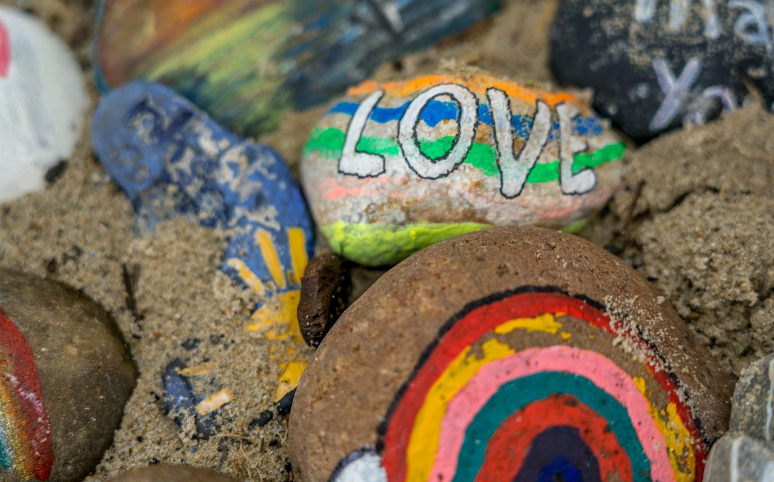 Coloridas rocas pintadas sobre la arena, incluida una con la palabra LOVE en letras gruesas con rayas arco iris y otra con un diseño arco iris.