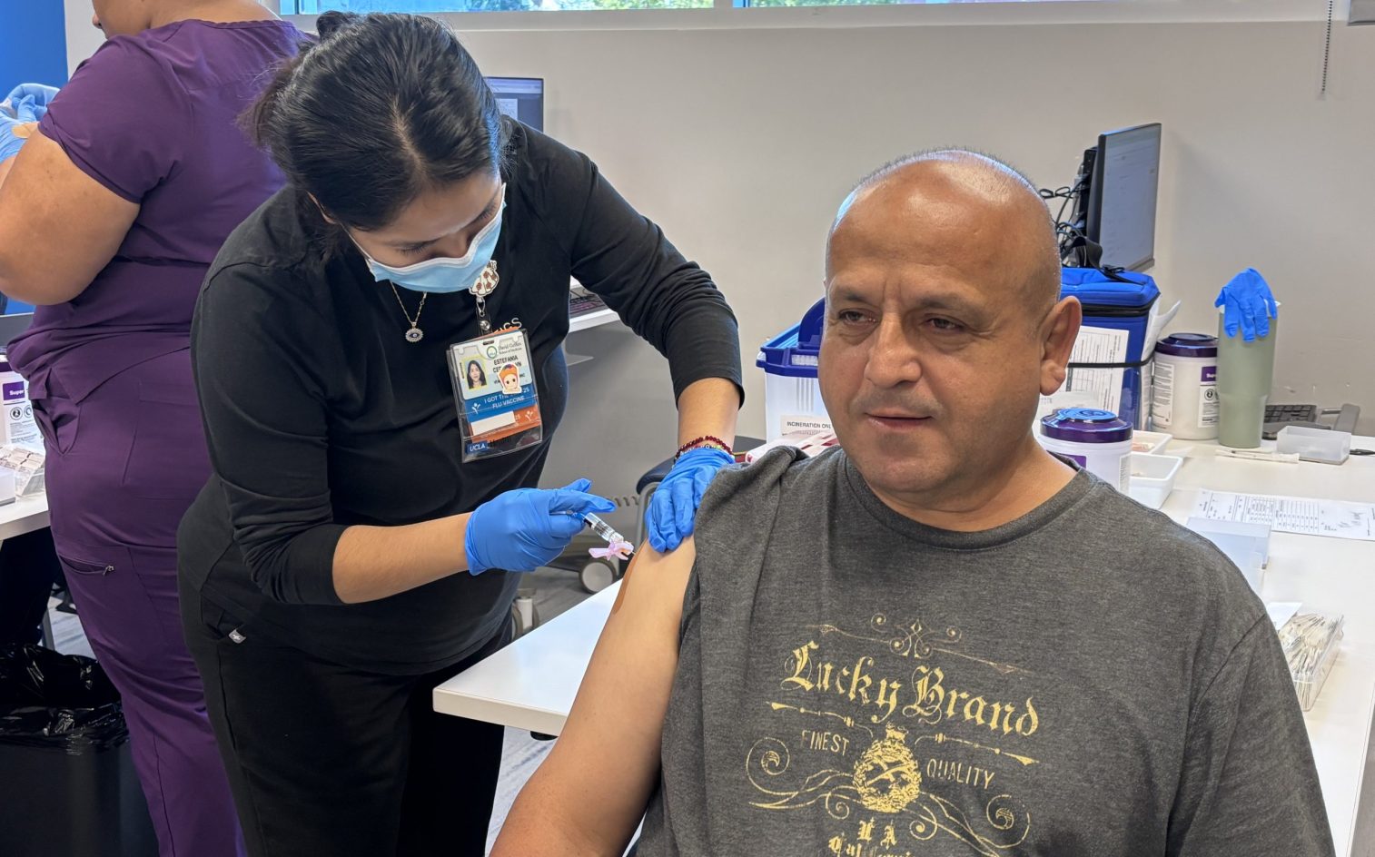 Un trabajador sanitario con bata y guantes administra una vacuna a un hombre con una camiseta de Lucky Brand, que está sentado con la manga remangada, en un entorno clínico con material médico visible en el mostrador. Un trabajador sanitario con bata y guantes administra una vacuna a un hombre con una camiseta de Lucky Brand, que está sentado con la manga remangada, en un entorno clínico con material médico visible en el mostrador.