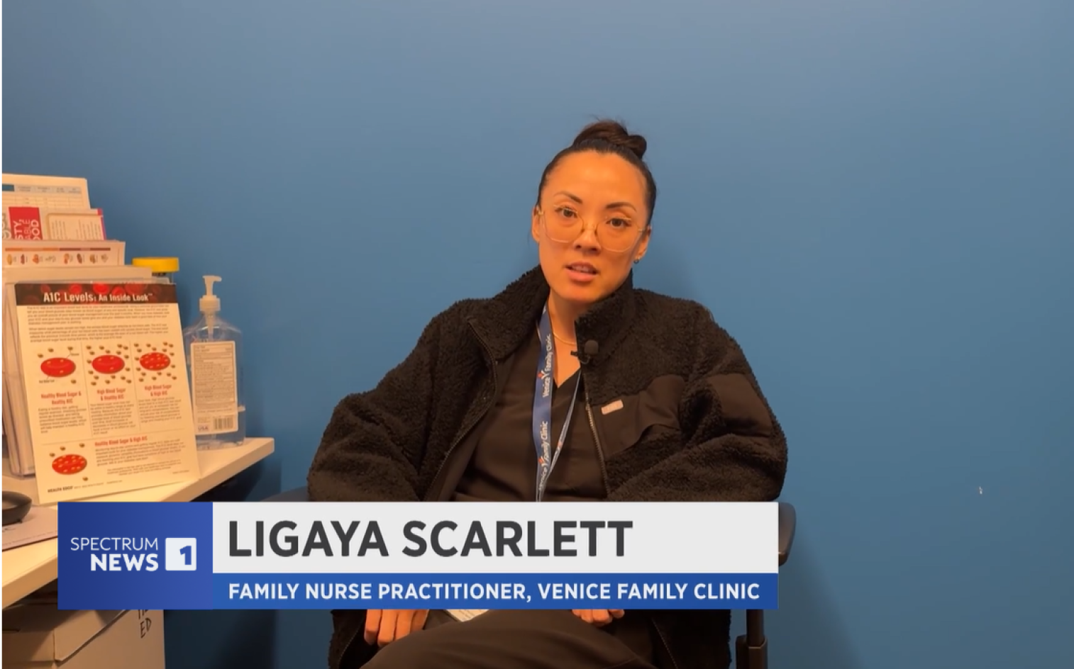 A woman wearing glasses and a black jacket sits in a clinic office with blue walls, speaking in an interview. Text on screen reads: Ligaya Scarlett, Family Nurse Practitioner, Venice Family Clinic and Spectrum News 1.