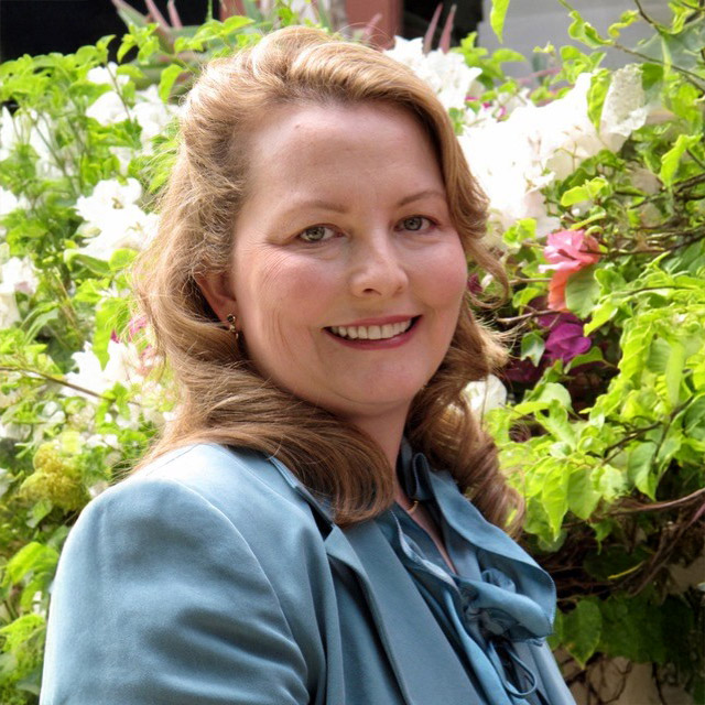 A Woman With Wavy Light Brown Hair, Wearing A Blue Blazer And Blouse, Smiles While Standing In Front Of A Background Of Green Leaves And White And Pink Flowers.