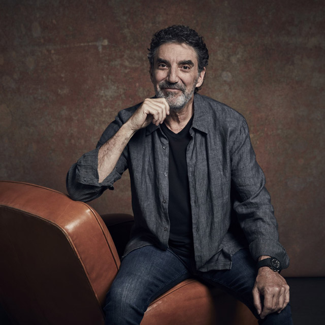 A Middle-aged Man With Curly Hair And A Beard Sits Casually On A Brown Leather Chair, Wearing A Dark Shirt, Jeans, And A Watch, Smiling Slightly With His Hand Resting On His Chin Against A Textured Brown Background.