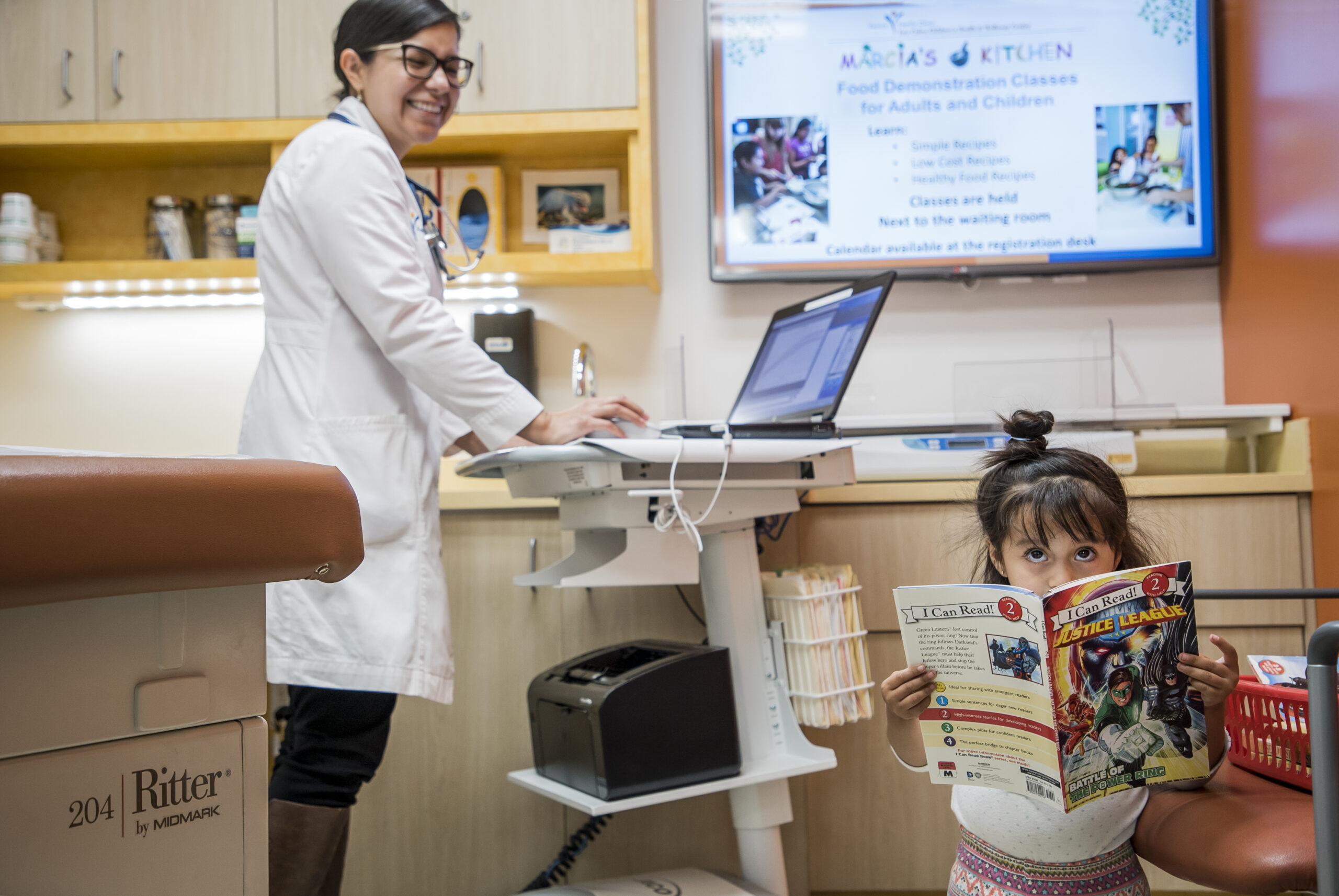 Una doctora sonríe mientras trabaja con un ordenador en una sala de reconocimiento médico. En primer plano, una niña está sentada en una mesa de exploración, sosteniendo y leyendo un cómic. En la pared de detrás se ve un póster de una clase de salud.
