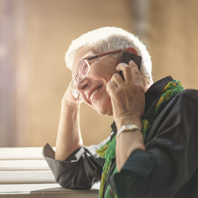 An Older Woman With Short Gray Hair And Glasses Smiles While Talking On A Smartphone, Sitting Near A Sunlit Window And Wearing A Dark Shirt With A Green Patterned Scarf.