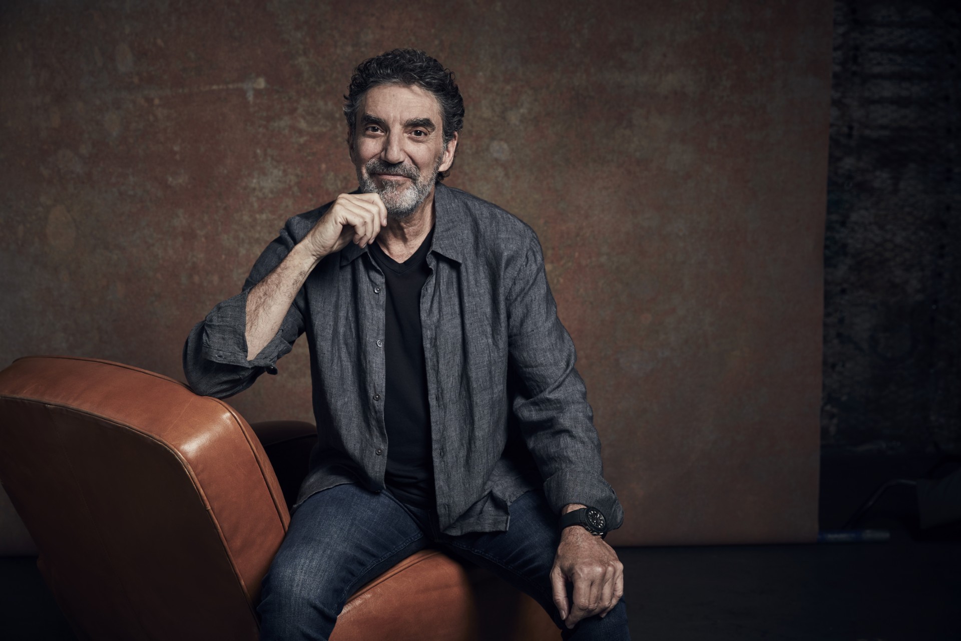 A Man With Gray Hair And A Beard Sits On A Brown Leather Chair, Wearing A Gray Shirt Over A Black T-shirt And Jeans. He Smiles Slightly And Rests His Chin On His Hand Against A Plain Brown Background. A Man With Gray Hair And A Beard Sits On A Brown Leather Chair, Wearing A Gray Shirt Over A Black T-shirt And Jeans. He Smiles Slightly And Rests His Chin On His Hand Against A Plain Brown Background.