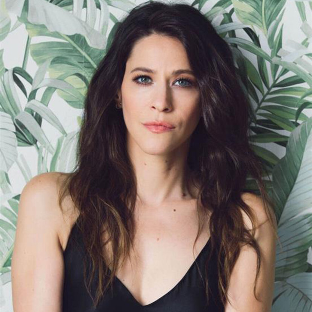 A Woman With Long, Wavy Dark Hair And Blue Eyes Stands In Front Of A Leafy, Tropical-patterned Background, Wearing A Black Sleeveless Top And Looking Seriously At The Camera.