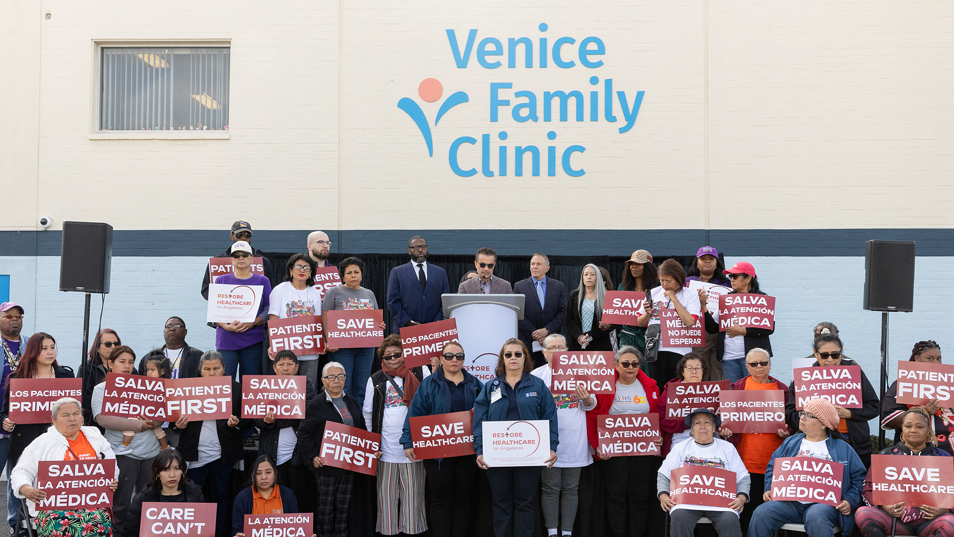 Un grupo heterogéneo de personas con pancartas que rezan "Salvemos la sanidad" y "Los pacientes primero" en inglés y español, reunidas en torno a un orador en un podio durante una concentración o protesta.