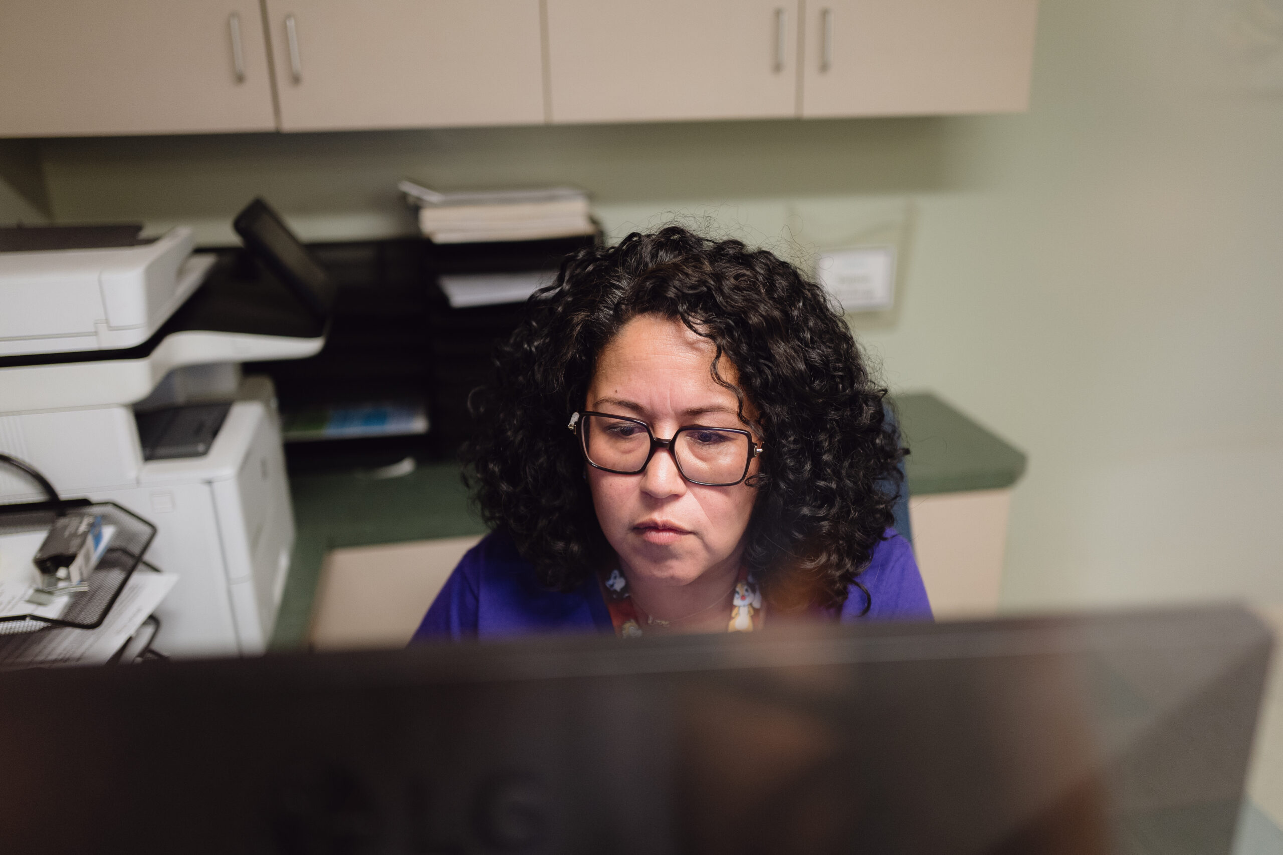 A Woman With Curly Hair And Glasses Works At A Computer In An Office, With Cabinets, Papers, And A Printer In The Background. A Woman With Curly Hair And Glasses Works At A Computer In An Office, With Cabinets, Papers, And A Printer In The Background.